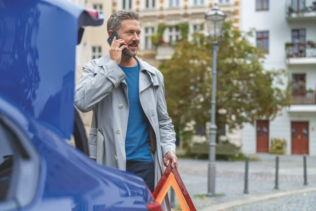 Kaltenbach Gruppe – Ein Mann in einem grauen Mantel steht an einem offenen Kofferraum, hält ein Warndreieck in der Hand und telefoniert über Serviceleistungen. Er wirkt besorgt und steht in einer Wohnstraße mit Gebäuden im Hintergrund. – BMW, MINI, KIA, BMW Motorrad & Honda Motorrad Partner in NRW