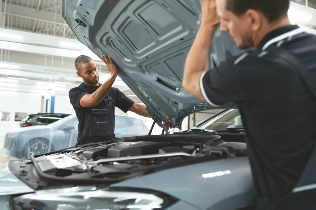 Kaltenbach Gruppe – Zwei Automechaniker in Uniform arbeiten in einer hell erleuchteten Werkstatt bei geöffneter Motorhaube zusammen und inspizieren den Motor und die Bauteile im Inneren, während sie über bevorstehende Serviceleistungen und Termine sprechen. – BMW, MINI, KIA, BMW Motorrad & Honda Motorrad Partner in NRW