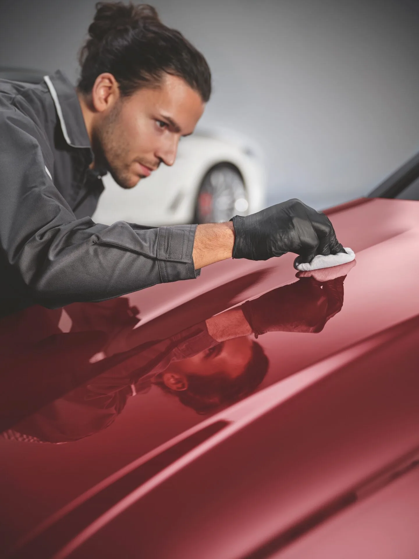 Kaltenbach Gruppe – Ein Mann in Handschuhen und Arbeitsuniform poliert sorgfältig die glänzende rote Motorhaube eines Autos, sein Spiegelbild ist auf der Oberfläche zu sehen. Im Hintergrund ist unscharf ein weißes Auto zu sehen. Professionelle Serviceleistungen sind nach Terminvereinbarung möglich. – BMW, MINI, KIA, BMW Motorrad & Honda Motorrad Partner in NRW