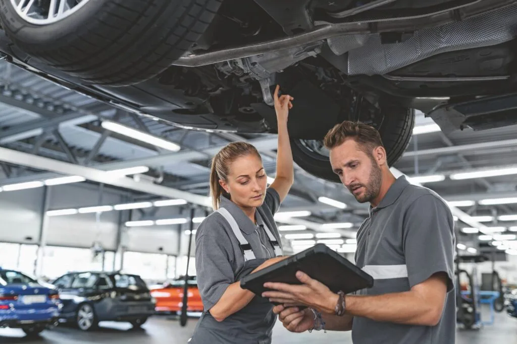 Kaltenbach Gruppe – Zwei Automechaniker in grauen Uniformen stehen unter einem angehobenen Auto. Einer zeigt auf den Reifen, der andere hält ein Tablet in der Hand. Beide scheinen konzentriert zu sein und besprechen Diagnosen in einer hellen Garage mit anderen Rädern und Fahrzeugen im Hintergrund. – BMW, MINI, KIA, BMW Motorrad & Honda Motorrad Partner in NRW