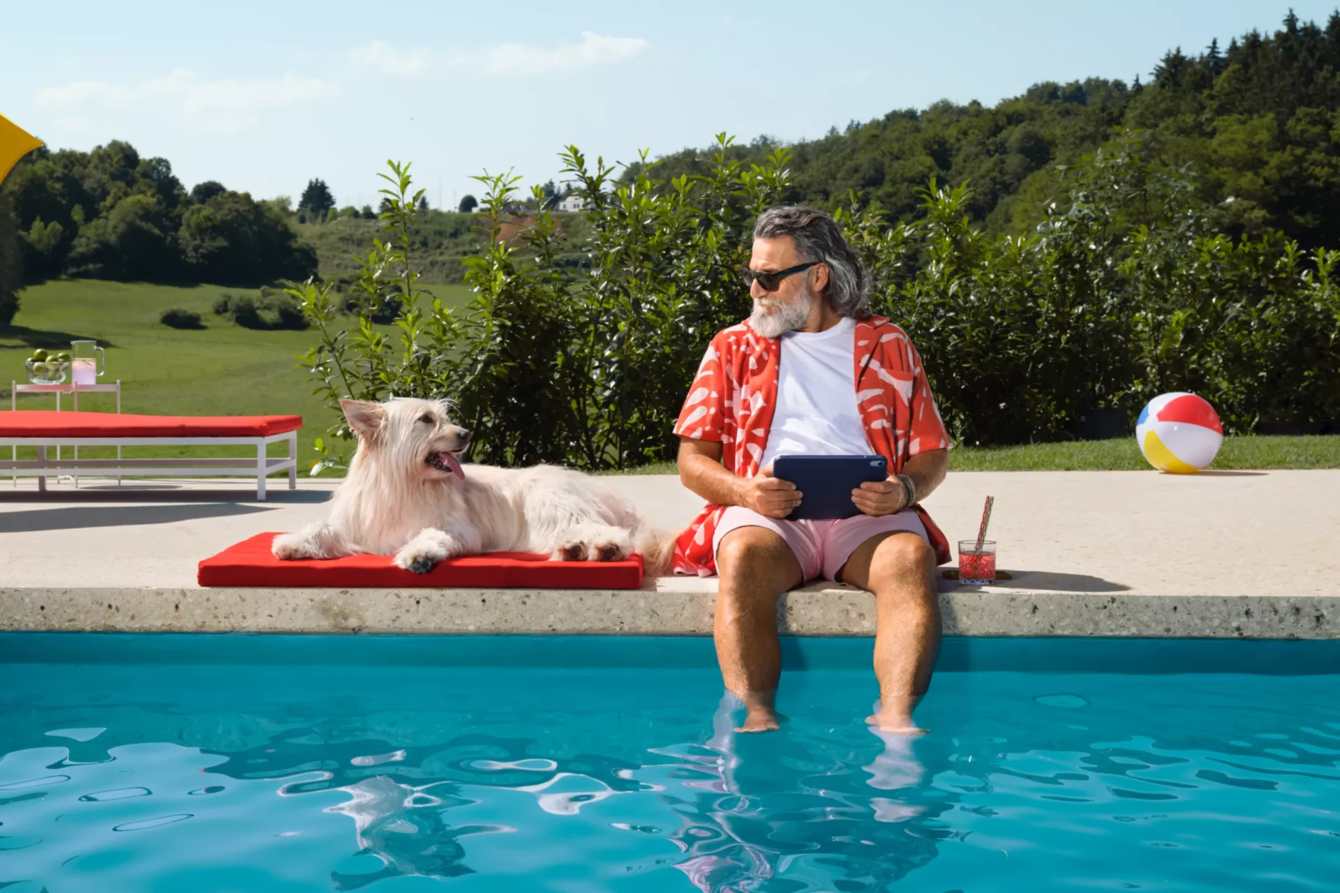 Kaltenbach Gruppe – Ein Mann mit rotem Hemd und Sonnenbrille sitzt an einem Pool, die Füße im Wasser, und erkundet Servicepakete auf seinem Tablet. Sein weißer Hund ruht in der Nähe auf einer roten Matte, mit einem Getränk und einem Strandball in der Nähe, umgeben von Bäumen und Hügeln. – BMW, MINI, KIA, BMW Motorrad & Honda Motorrad Partner in NRW