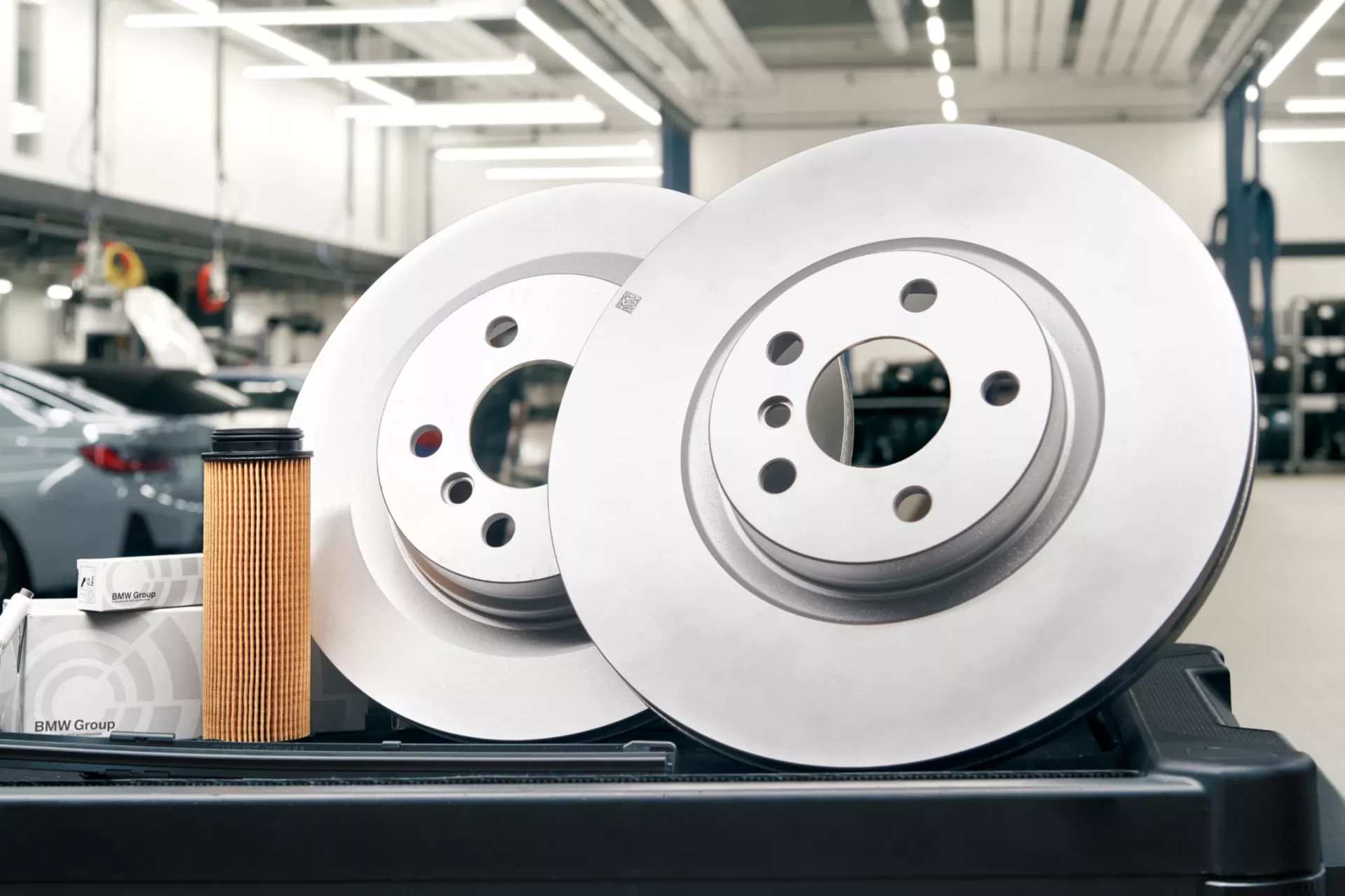 Kaltenbach Gruppe – Zwei glänzende, neue Bremsscheiben, ein Ölfilter und eine BMW Group Teilebox liegen auf einer Werkbank in einer hell erleuchteten Kfz-Werkstatt - perfekte Voraussetzungen für umfangreiche Servicepakete. – BMW, MINI, KIA, BMW Motorrad & Honda Motorrad Partner in NRW