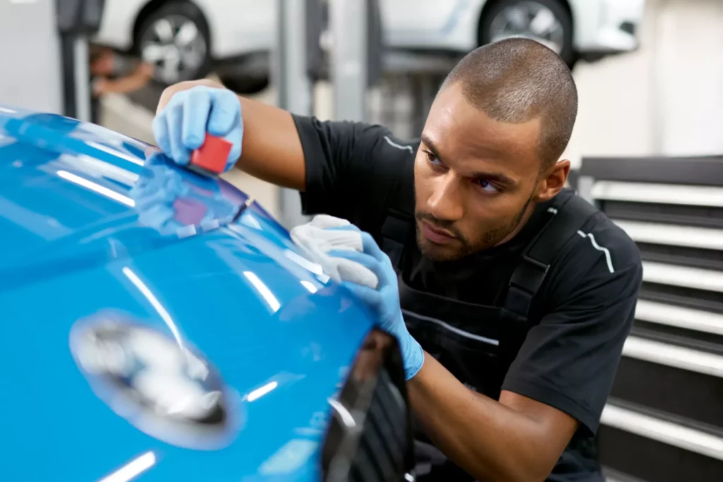Kaltenbach Gruppe – Ein zielstrebiger Mann mit Handschuhen benutzt ein rotes Werkzeug, um die leuchtend blaue Motorhaube eines Autos in einer Autowerkstatt sorgfältig zu polieren oder mit einer Schutzfolie zu versehen, um eines ihrer hochwertigen Servicepakete zu präsentieren. – BMW, MINI, KIA, BMW Motorrad & Honda Motorrad Partner in NRW