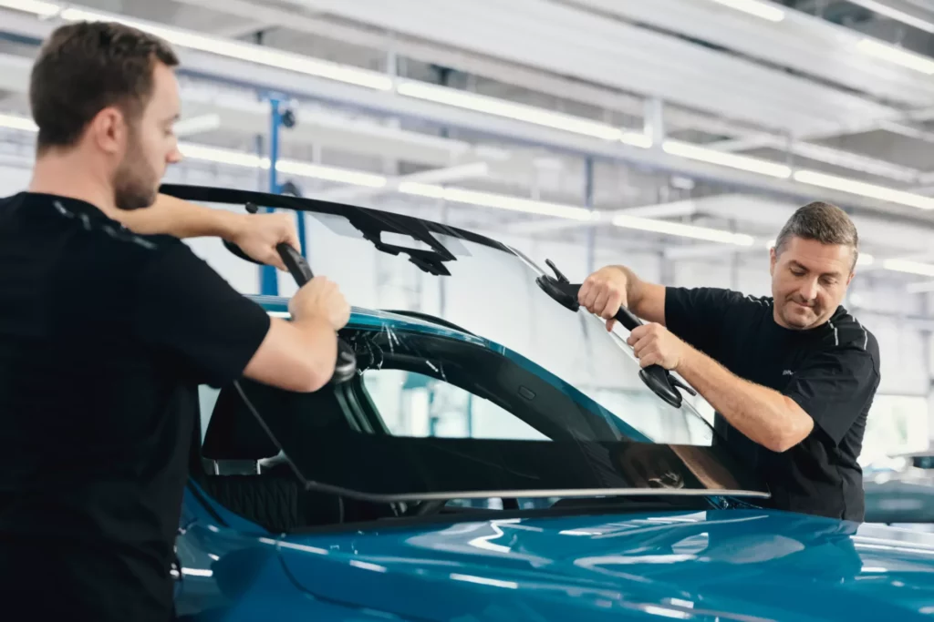 Kaltenbach Gruppe – Zwei Männer in schwarzen Hemden montieren bzw. tauschen in einer gut beleuchteten Kfz-Werkstatt eine Windschutzscheibe an einem blauen Auto aus und sorgen im Rahmen ihrer umfassenden Servicepakete für ein qualitativ hochwertiges Ergebnis. – BMW, MINI, KIA, BMW Motorrad & Honda Motorrad Partner in NRW