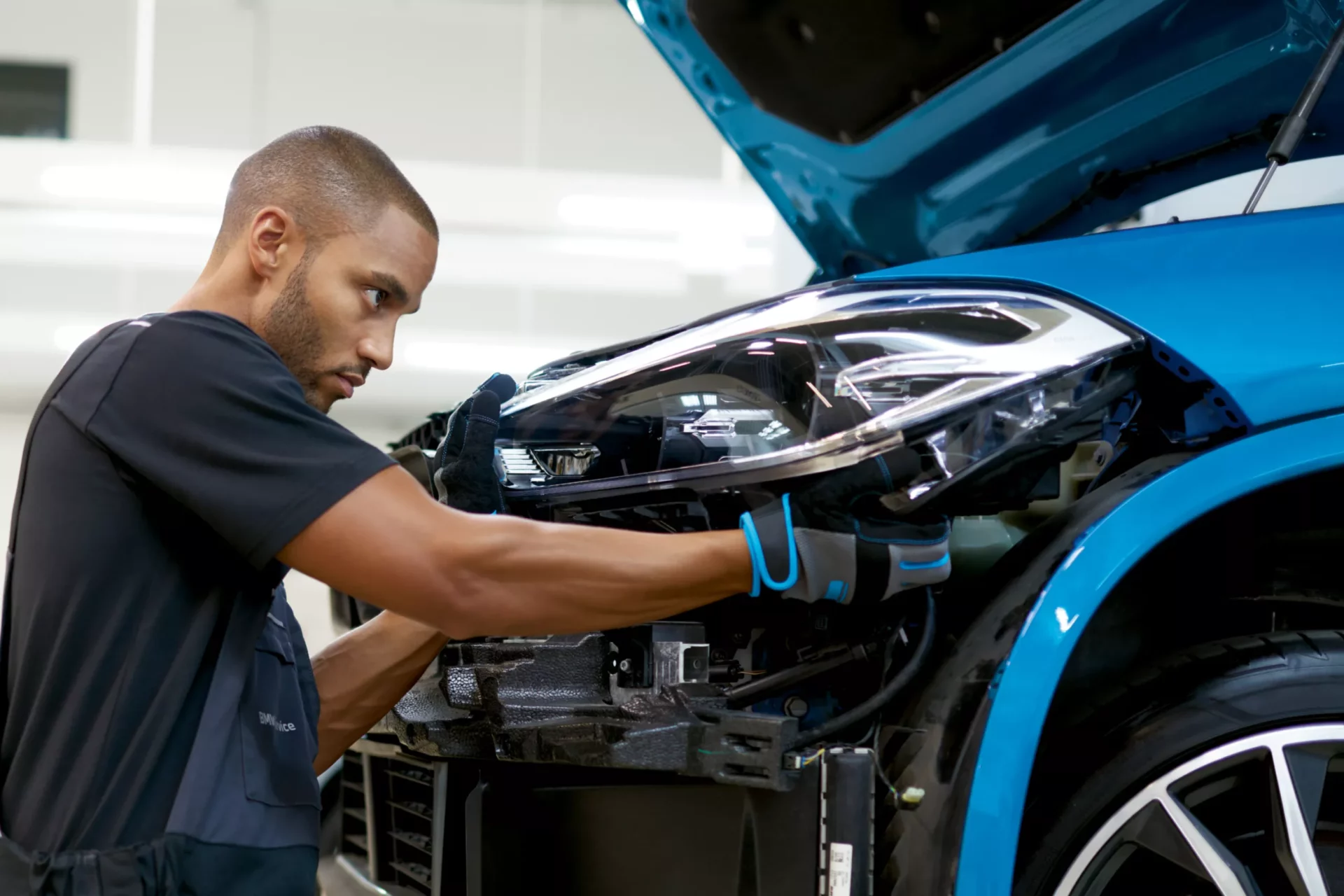 Kaltenbach Gruppe – Ein Mann mit Handschuhen arbeitet konzentriert an der Scheinwerfereinheit eines blauen Autos mit geöffneter Motorhaube und nimmt Reparaturen oder Einstellungen in einer gut beleuchteten Werkstatt vor, die verschiedene Servicepakete für die Fahrzeugwartung anbietet. – BMW, MINI, KIA, BMW Motorrad & Honda Motorrad Partner in NRW