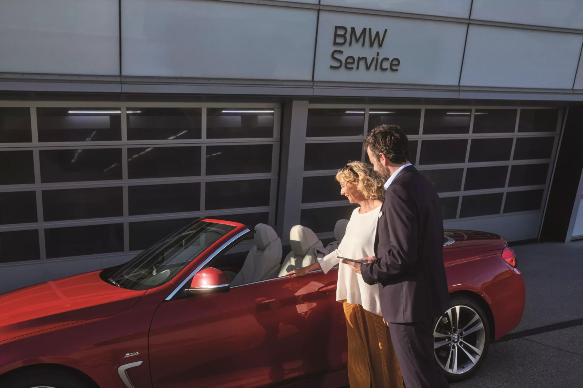 Kaltenbach Gruppe – Ein Mann und eine Frau stehen neben einem roten BMW Cabrio, das vor einem Gebäude mit der Aufschrift BMW Service geparkt ist. Die sonnenbeschienene Szene lässt vermuten, dass sie sich über Servicepakete unterhalten, während sie in ein Gespräch vertieft sind. – BMW, MINI, KIA, BMW Motorrad & Honda Motorrad Partner in NRW
