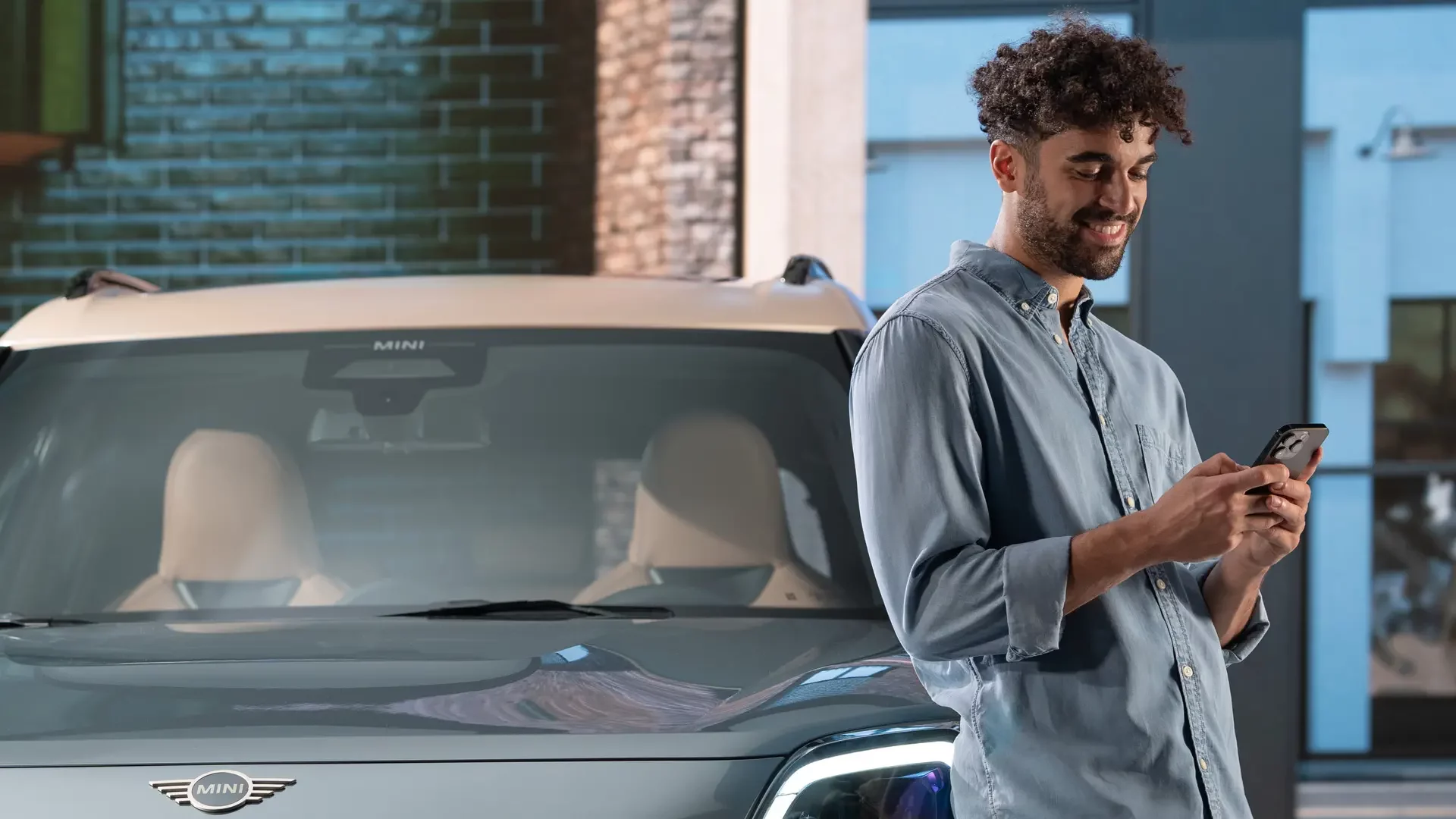 Kaltenbach Gruppe – Ein Mann mit lockigem Haar steht lächelnd und mit Blick auf sein Handy vor einem geparkten MINI Auto in einem Innenraum. Die Windschutzscheibe und das Logo des Autos sind zu sehen, mit Ziegelsteinen und blauen Wänden im Hintergrund, was darauf hindeutet, dass er MINI Servicepakete erkunden möchte. – BMW, MINI, KIA, BMW Motorrad & Honda Motorrad Partner in NRW