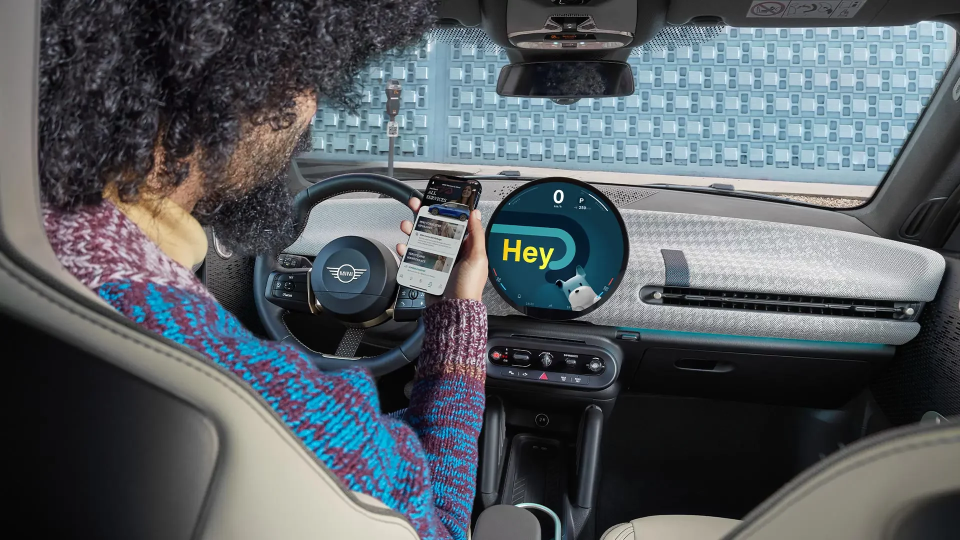 Kaltenbach Gruppe – Eine Person sitzt auf dem Fahrersitz eines modernen Autos und hält ein Smartphone in der Hand. Das digitale Armaturenbrett zeigt "Hey" an und weist auf Konnektivitätsfunktionen und Servicepakete hin. Der stilvolle Innenraum zeichnet sich durch gemusterte Sitze und ein einzigartiges Armaturenbrett-Design aus. – BMW, MINI, KIA, BMW Motorrad & Honda Motorrad Partner in NRW