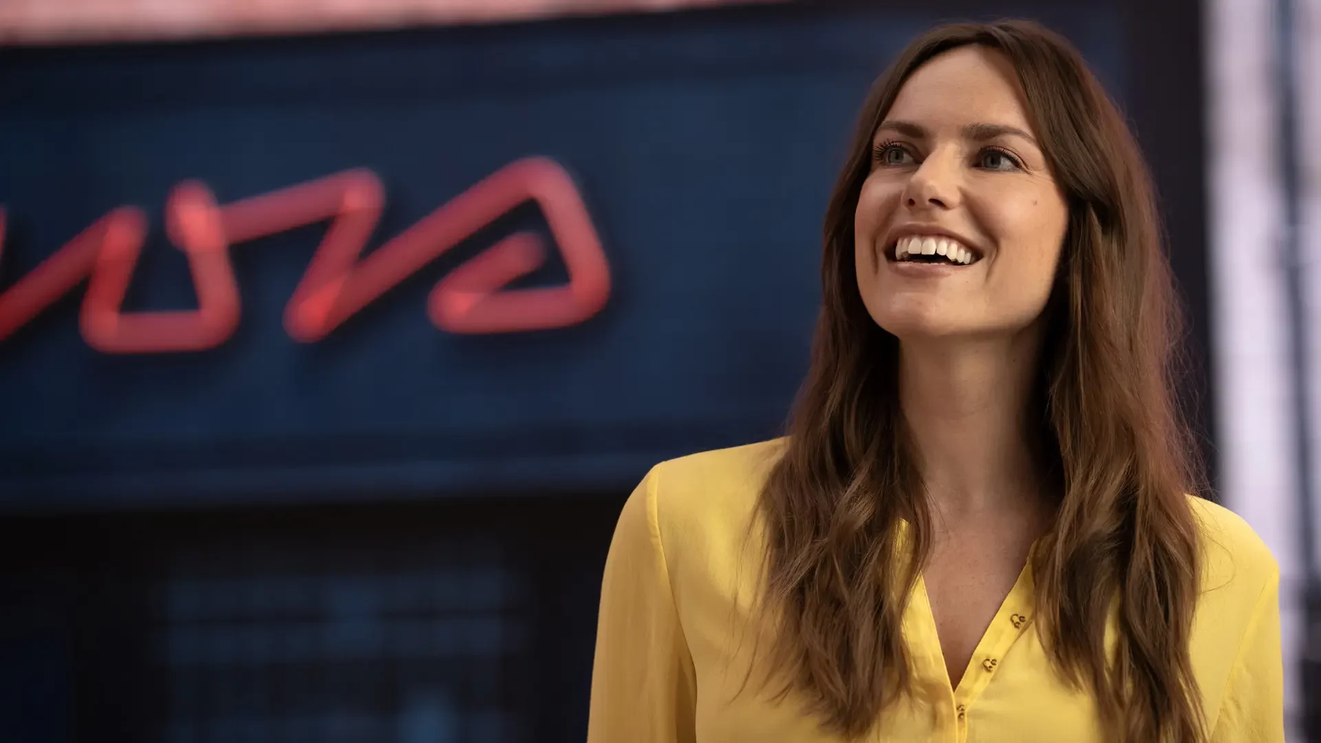 Kaltenbach Gruppe – Eine Frau mit langen braunen Haaren und gelber Bluse lächelt, während sie im Freien vor einem Gebäude mit verschwommener roter Neonreklame steht, die die pulsierende Energie widerspiegelt, die oft mit modernen Servicepaketen assoziiert wird. – BMW, MINI, KIA, BMW Motorrad & Honda Motorrad Partner in NRW