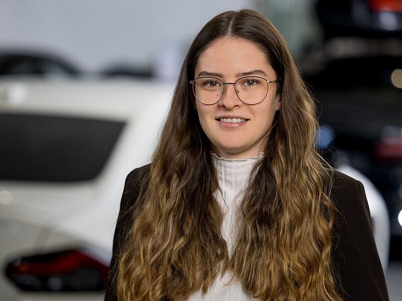 Kaltenbach Gruppe – Eine junge Frau mit langen, gewellten braunen Haaren, die eine Brille, eine weiße Bluse und einen dunklen Blazer trägt, steht in einer Halle in Kaltenbach Overath vor verschwommenen Autos. Sie blickt in die Kamera und lächelt leicht. – BMW, MINI, KIA, BMW Motorrad & Honda Motorrad Partner in NRW