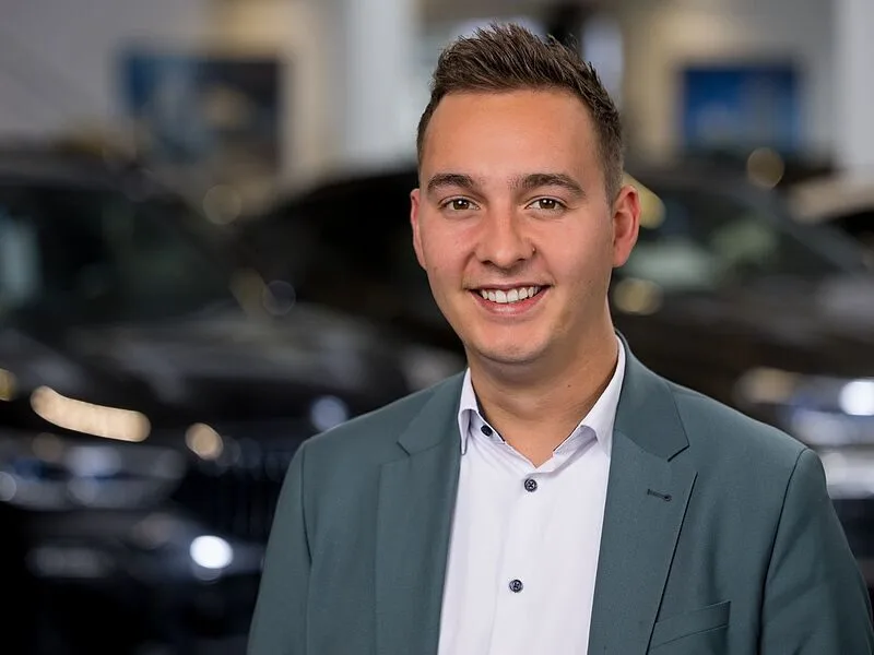 Kaltenbach Gruppe – Ein junger Mann in Anzugjacke und weißem Hemd lächelt in die Kamera, als er im hell erleuchteten Ausstellungsraum von Kaltenbach Remscheid vor dunkelfarbigen Autos steht. – BMW, MINI, KIA, BMW Motorrad & Honda Motorrad Partner in NRW
