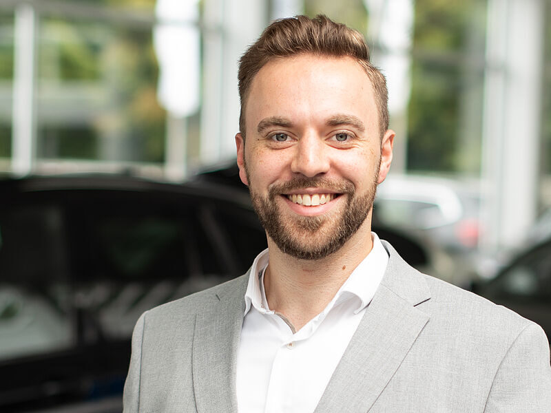 Kaltenbach Gruppe – Ein lächelnder Mann mit kurzen braunen Haaren und Bart, der eine hellgraue Anzugsjacke und ein weißes Hemd trägt, steht in einem Innenraum der Kaltenbach-Zentrale, mit unscharfen Autos und Fenstern im Hintergrund. – BMW, MINI, KIA, BMW Motorrad & Honda Motorrad Partner in NRW