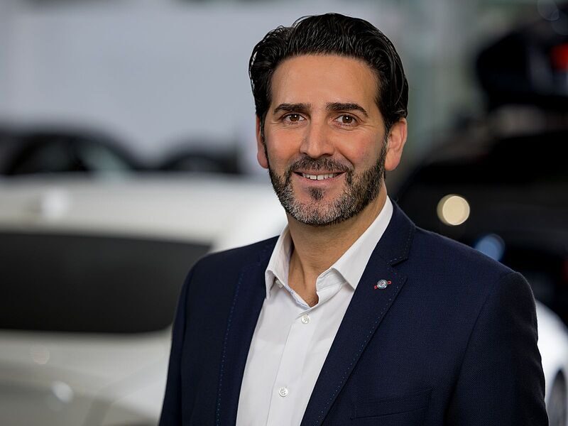 Kaltenbach Gruppe – Ein Mann mit dunklem Haar und Bart, der einen marineblauen Blazer und ein weißes Hemd trägt, lächelt in einer modernen Inneneinrichtung in Kaltenbach Waldbröl in die Kamera, mit unscharfen Autos im Hintergrund. – BMW, MINI, KIA, BMW Motorrad & Honda Motorrad Partner in NRW