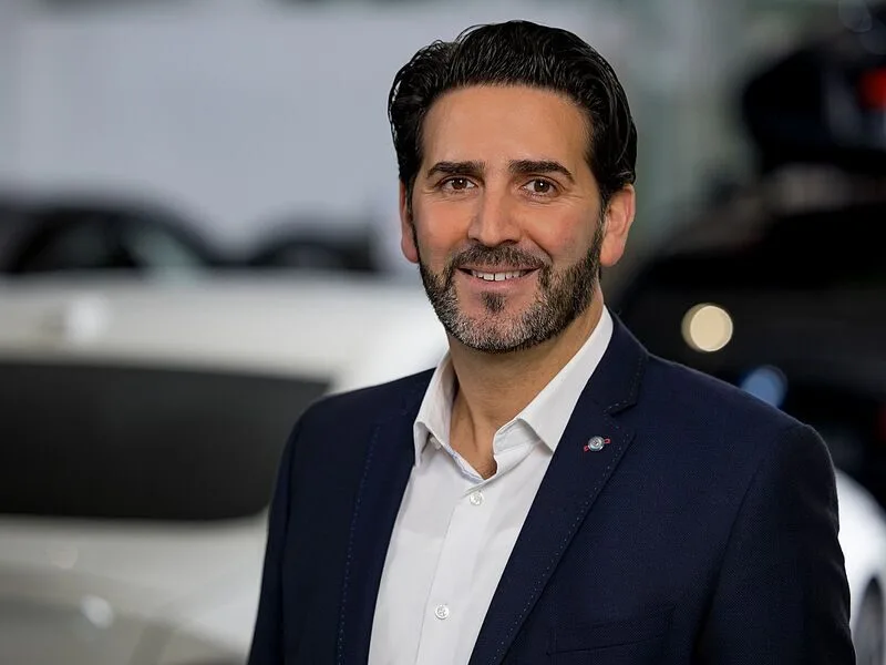 Kaltenbach Gruppe – Ein Mann mit dunklem Haar und Bart, der einen marineblauen Blazer und ein weißes Hemd trägt, lächelt in einer modernen Inneneinrichtung in Kaltenbach Waldbröl in die Kamera, mit unscharfen Autos im Hintergrund. – BMW, MINI, KIA, BMW Motorrad & Honda Motorrad Partner in NRW