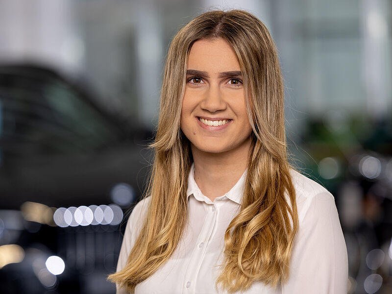 Kaltenbach Gruppe – Eine junge Frau mit langen blonden Haaren, die ein weißes Button-up-Hemd trägt, lächelt in die Kamera. Der sanft beleuchtete, unscharfe Innenhintergrund deutet auf einen einladenden Raum in Kaltenbach Overath hin. – BMW, MINI, KIA, BMW Motorrad & Honda Motorrad Partner in NRW
