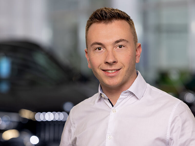 Kaltenbach Gruppe – Ein junger Mann mit kurzen hellbraunen Haaren, der ein helles Button-up-Hemd trägt, lächelt in einem Innenraum in Kaltenbach Overath in die Kamera, mit unscharfen Autos im Hintergrund. – BMW, MINI, KIA, BMW Motorrad & Honda Motorrad Partner in NRW