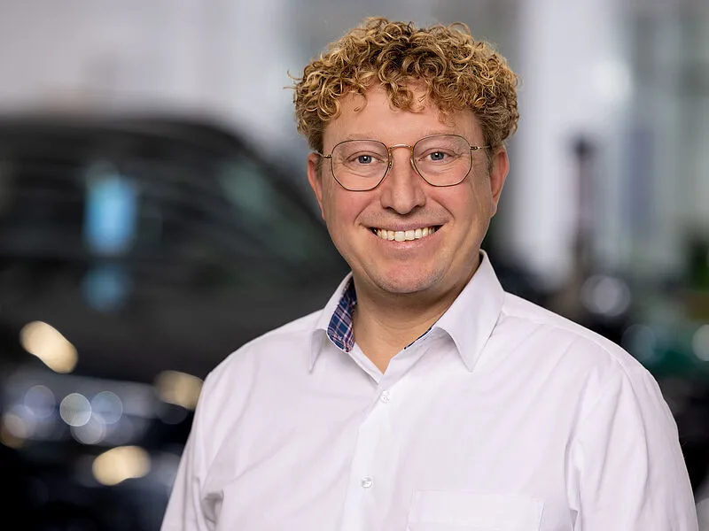 Kaltenbach Gruppe – Ein lächelnder Mann mit lockigem blondem Haar und Brille, der ein weißes Hemd trägt, steht vor einem unscharfen Innenhintergrund in Kaltenbach Wermelskirchen. – BMW, MINI, KIA, BMW Motorrad & Honda Motorrad Partner in NRW