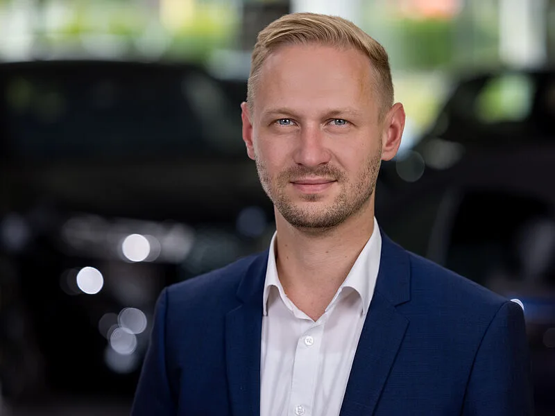 Kaltenbach Gruppe – Ein Mann mit kurzen blonden Haaren und gestutztem Bart, blauer Anzugsjacke und weißem Hemd steht in einem Innenraum in Kaltenbach Olpe, mit unscharfen Autos und Lichtern im Hintergrund. – BMW, MINI, KIA, BMW Motorrad & Honda Motorrad Partner in NRW