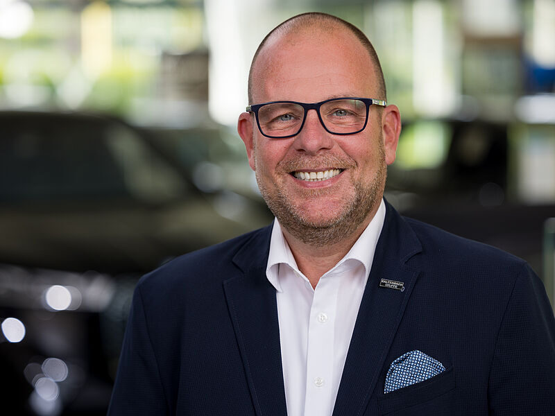 Kaltenbach Gruppe – Ein lächelnder Mann mit Brille und gestutztem Bart, der eine dunkle Anzugsjacke, ein weißes Hemd und ein Einstecktuch trägt, steht in einem Innenraum der Kaltenbach-Zentrale vor einem sanft verschwommenen Hintergrund. – BMW, MINI, KIA, BMW Motorrad & Honda Motorrad Partner in NRW