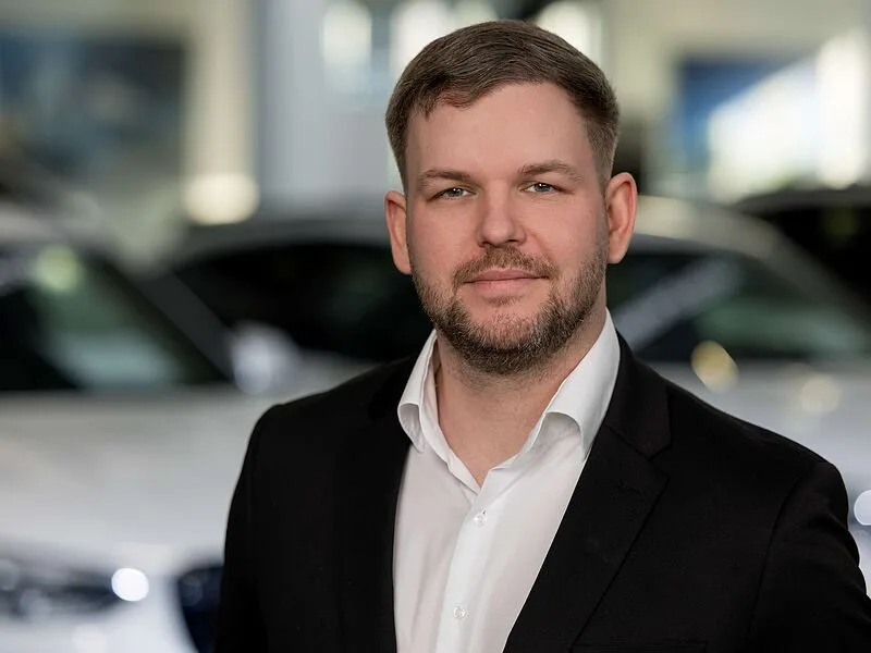 Kaltenbach Gruppe – Ein Mann mit kurzen Haaren und Bart, der ein weißes Hemd und eine schwarze Anzugsjacke trägt, steht in einer Halle in Kaltenbach-Remscheid vor verschwommenen Autos und blickt mit einem leichten Lächeln in die Kamera. – BMW, MINI, KIA, BMW Motorrad & Honda Motorrad Partner in NRW
