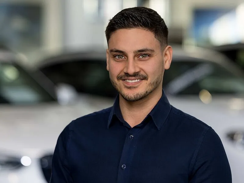 Kaltenbach Gruppe – Ein lächelnder Mann mit kurzen dunklen Haaren und einem ordentlich gestutzten Bart, der ein marineblaues Hemd trägt, steht in einer Halle in Kaltenbach Werdohl, mit unscharfen weißen Autos im Hintergrund. – BMW, MINI, KIA, BMW Motorrad & Honda Motorrad Partner in NRW