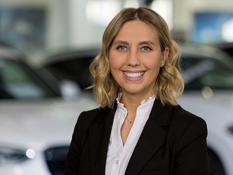 Kaltenbach Gruppe – Eine lächelnde Frau mit blondem, gewelltem Haar, die einen schwarzen Blazer und eine weiße Bluse trägt, steht in einem Haus in Kaltenbach-Remscheid vor verschwommenen weißen Autos. – BMW, MINI, KIA, BMW Motorrad & Honda Motorrad Partner in NRW