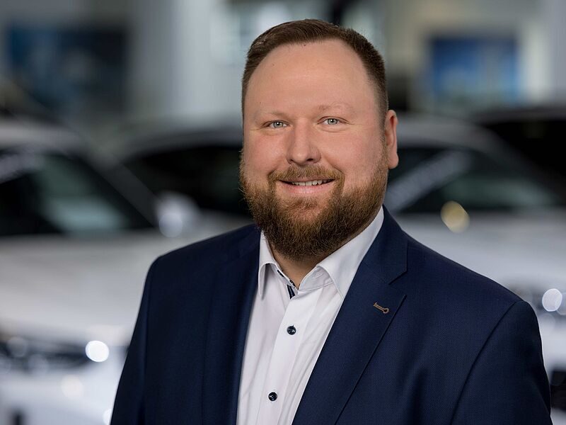 Kaltenbach Gruppe – Ein lächelnder Mann mit Bart, der einen marineblauen Anzug und ein weißes Hemd trägt, steht in einem Innenraum vor verschwommenen weißen Autos in Kaltenbach Werdohl. – BMW, MINI, KIA, BMW Motorrad & Honda Motorrad Partner in NRW