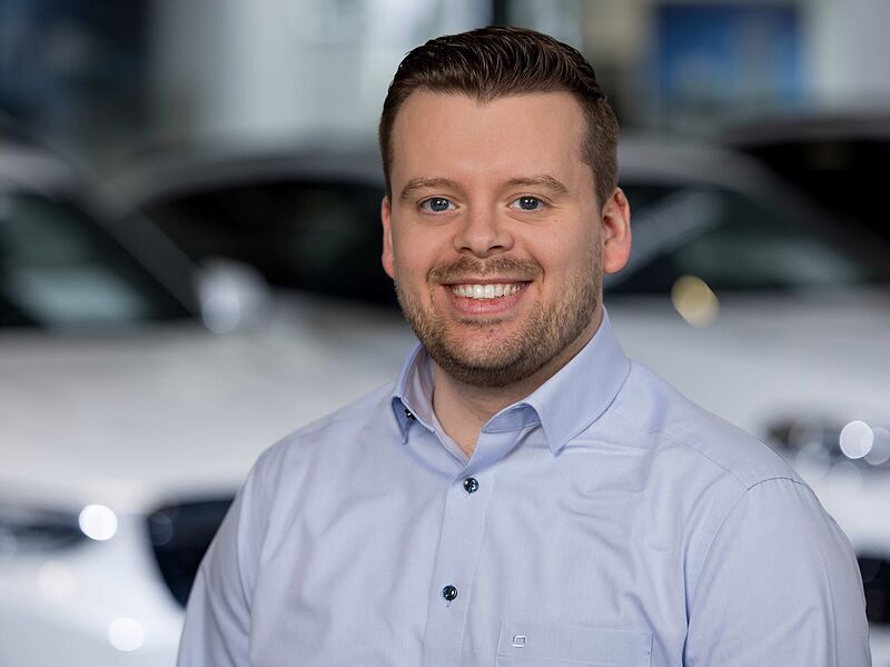 Kaltenbach Gruppe – Ein Mann mit kurzen braunen Haaren und Bart, der ein hellblaues Button-up-Hemd trägt, lächelt, während er im Ausstellungsraum von Kaltenbach Remscheid steht, mit verschwommenen weißen Autos im Hintergrund. – BMW, MINI, KIA, BMW Motorrad & Honda Motorrad Partner in NRW