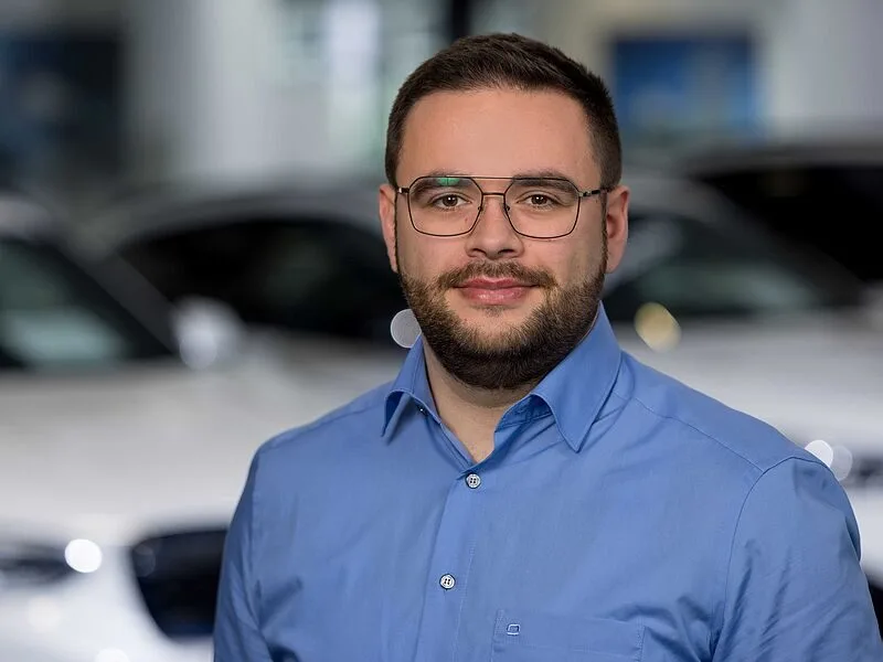 Kaltenbach Gruppe – Ein Mann mit kurzen braunen Haaren, Bart und Brille, der ein blaues Hemd zum Knöpfen trägt, steht in einem Innenraum vor verschwommenen weißen Autos in Kaltenbach-Remscheid. – BMW, MINI, KIA, BMW Motorrad & Honda Motorrad Partner in NRW