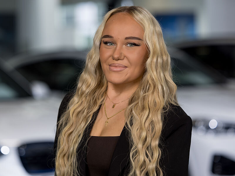 Kaltenbach Gruppe – Eine Frau mit langem, gewelltem blondem Haar lächelt in die Kamera. Sie trägt einen schwarzen Blazer, ein dunkles Top und mehrere goldene Halsketten. Im Hintergrund sind verschwommene weiße Autos zu sehen, die auf einen geschäftigen Tag in Kaltenbach-Werdohl hindeuten. – BMW, MINI, KIA, BMW Motorrad & Honda Motorrad Partner in NRW