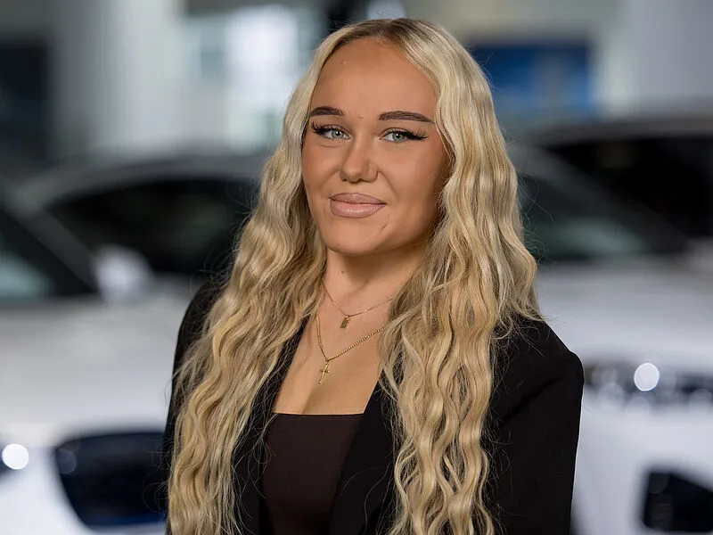 Kaltenbach Gruppe – Eine Frau mit langem, gewelltem blondem Haar lächelt in die Kamera. Sie trägt einen schwarzen Blazer, ein dunkles Top und mehrere goldene Halsketten. Im Hintergrund sind verschwommene weiße Autos zu sehen, die auf einen geschäftigen Tag in Kaltenbach-Werdohl hindeuten. – BMW, MINI, KIA, BMW Motorrad & Honda Motorrad Partner in NRW