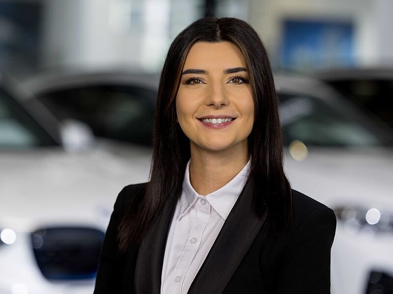 Kaltenbach Gruppe – Eine Frau mit langen dunklen Haaren, die ein weißes Hemd und einen schwarzen Blazer trägt, lächelt, während sie in einer gut beleuchteten Halle in Kaltenbach Olpe vor verschwommenen weißen Autos steht. – BMW, MINI, KIA, BMW Motorrad & Honda Motorrad Partner in NRW