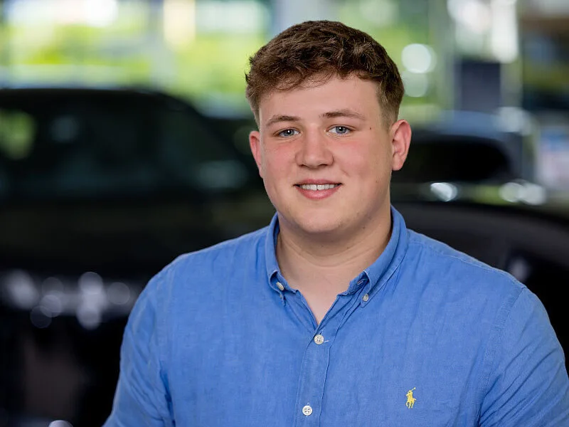 Kaltenbach Gruppe – Ein junger Mann mit kurzen braunen Haaren und blauem Hemd lächelt, während er in einem Haus in Kaltenbach Overath steht. Im Hintergrund sind unscharfe Autos zu sehen. – BMW, MINI, KIA, BMW Motorrad & Honda Motorrad Partner in NRW