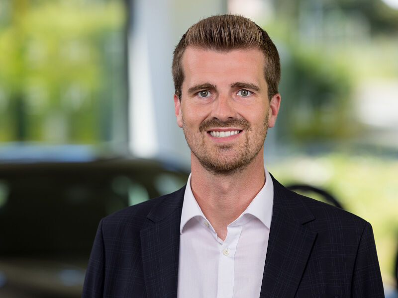 Kaltenbach Gruppe – Ein Mann mit kurzen braunen Haaren und gestutztem Bart, der eine dunkle Anzugsjacke und ein weißes Hemd trägt, steht lächelnd vor dem unscharfen, hellen Hintergrund der Kaltenbach Zentrale mit grünen und gelben Farbtönen. – BMW, MINI, KIA, BMW Motorrad & Honda Motorrad Partner in NRW