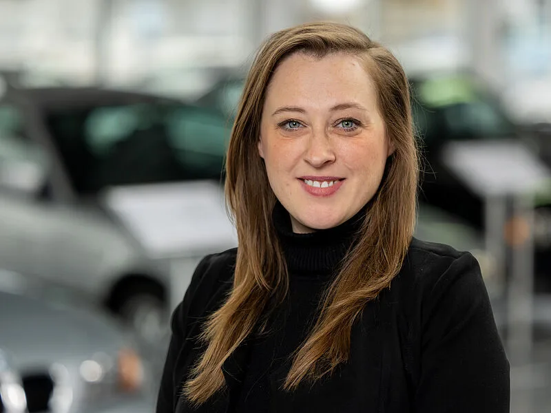 Kaltenbach Gruppe – Eine Frau mit langen hellbraunen Haaren, die einen schwarzen Rollkragenpullover und einen Blazer trägt, lächelt im Autohaus Kaltenbach Wipperfürth in die Kamera, während die Fahrzeuge im Hintergrund verschwimmen. – BMW, MINI, KIA, BMW Motorrad & Honda Motorrad Partner in NRW