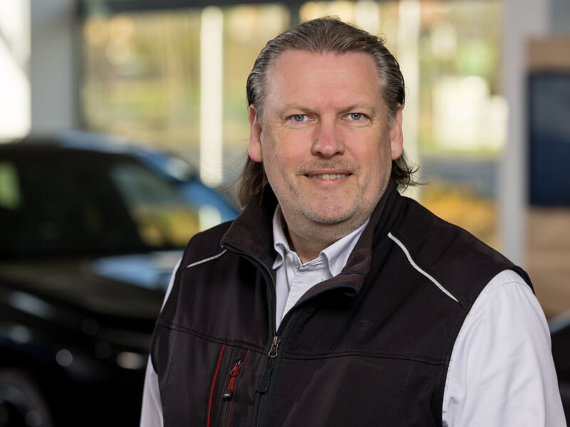 Kaltenbach Gruppe – Ein Mann mittleren Alters mit langen Haaren und gestutztem Bart, der ein weißes Hemd und eine dunkle Weste trägt, lächelt in einem Haus in Kaltenbach Overath in die Kamera, im Hintergrund ist ein unscharfes Auto zu sehen. – BMW, MINI, KIA, BMW Motorrad & Honda Motorrad Partner in NRW