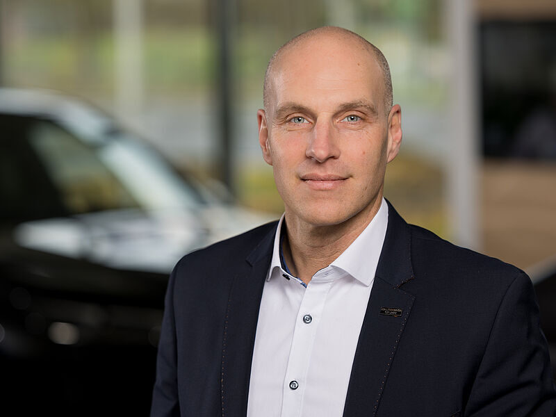 Kaltenbach Gruppe – Ein glatzköpfiger Mann in einem dunklen Anzug und weißem Hemd steht in einem Innenraum der Kaltenbach Zentrale und blickt mit neutralem Blick in die Kamera. Im Hintergrund sind ein verschwommenes Auto und große Fenster zu sehen. – BMW, MINI, KIA, BMW Motorrad & Honda Motorrad Partner in NRW