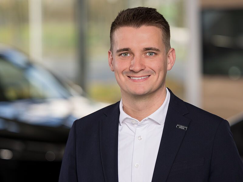 Kaltenbach Gruppe – Ein Mann in einem marineblauen Blazer und weißem Hemd lächelt in einer hellen Halle in der Kaltenbach Zentrale in die Kamera, während im Hintergrund Autos und Fenster verschwimmen. – BMW, MINI, KIA, BMW Motorrad & Honda Motorrad Partner in NRW