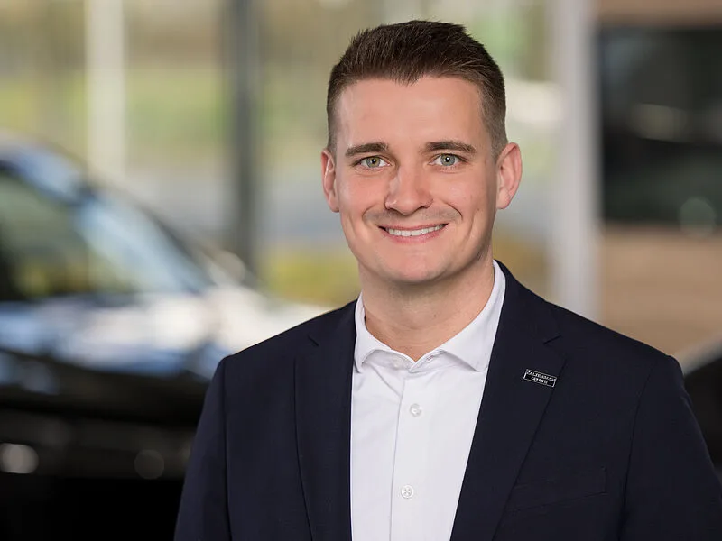Kaltenbach Gruppe – Ein Mann in einem marineblauen Blazer und weißem Hemd lächelt in einer hellen Halle in der Kaltenbach Zentrale in die Kamera, während im Hintergrund Autos und Fenster verschwimmen. – BMW, MINI, KIA, BMW Motorrad & Honda Motorrad Partner in NRW