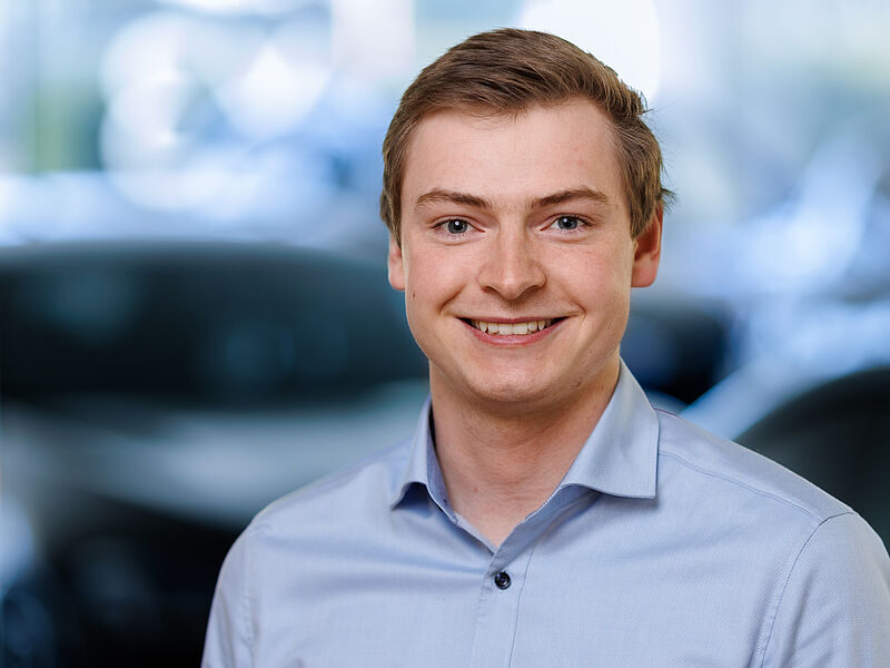 Kaltenbach Gruppe – Ein junger Mann mit kurzen hellbraunen Haaren lächelt in die Kamera. Er trägt ein hellblaues Hemd mit Kragen und steht vor einem unscharfen Hintergrund, vielleicht in der Nähe von Kaltenbach Waldbröl, mit dunklen und hellen Tönen. – BMW, MINI, KIA, BMW Motorrad & Honda Motorrad Partner in NRW