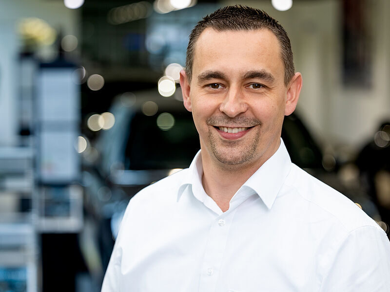 Kaltenbach Gruppe – Ein Mann mit kurzen dunklen Haaren und gestutztem Bart lächelt, während er ein weißes Hemd mit Kragen trägt. Er steht in einem Innenraum des Ausstellungsraums von Kaltenbach Overath mit einem unscharfen Bürohintergrund. – BMW, MINI, KIA, BMW Motorrad & Honda Motorrad Partner in NRW