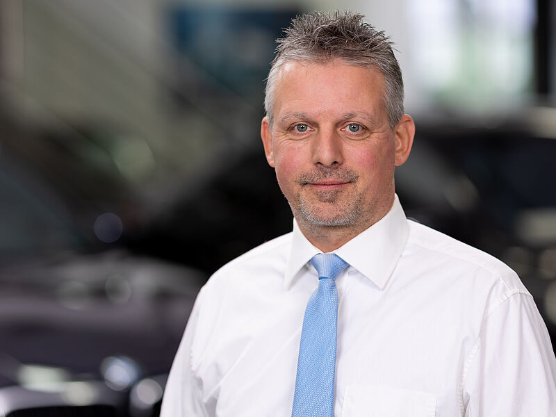 Kaltenbach Gruppe – Ein Mann mittleren Alters mit kurzen grauen Haaren, weißem Hemd und hellblauer Krawatte, steht in einem Haus und lächelt leicht in die Kamera. Der unscharfe Hintergrund suggeriert eine berufliche oder bürotechnische Umgebung in Kaltenbach Remscheid. – BMW, MINI, KIA, BMW Motorrad & Honda Motorrad Partner in NRW
