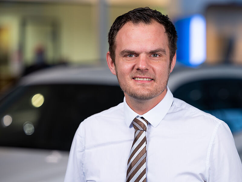 Kaltenbach Gruppe – Ein Mann mit weißem Hemd und gestreifter Krawatte lächelt in die Kamera. Er steht in einem Innenraum von Kaltenbach Werdohl, mit verschwommenen Autos und Ausstellungsräumen im Hintergrund. – BMW, MINI, KIA, BMW Motorrad & Honda Motorrad Partner in NRW