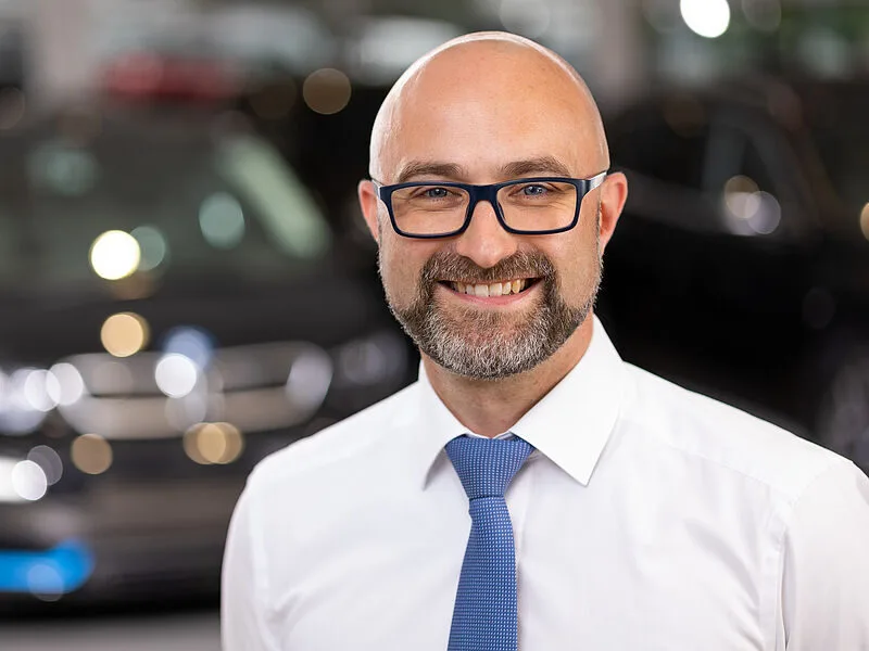 Kaltenbach Gruppe – Ein lächelnder Mann mit Glatze, Brille, Bart und blauer Krawatte steht in einer Halle in Kaltenbach Werdohl vor einem unscharfen Hintergrund mit Autos. – BMW, MINI, KIA, BMW Motorrad & Honda Motorrad Partner in NRW