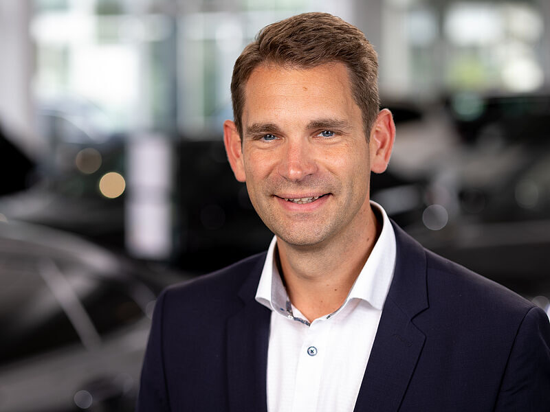 Kaltenbach Gruppe – Ein Mann in einer marineblauen Anzugsjacke und einem weißen Hemd lächelt in die Kamera in einem hellen, modernen Ausstellungsraum der Kaltenbach-Zentrale, mit verschwommenen Autos und großen Fenstern im Hintergrund. – BMW, MINI, KIA, BMW Motorrad & Honda Motorrad Partner in NRW