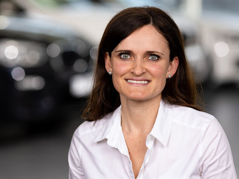 Kaltenbach Gruppe – Eine Frau mit langen braunen Haaren, die ein weißes Hemd mit Knöpfen trägt, lächelt in die Kamera. Der verschwommene Hintergrund mit unscharfen Autos lässt vermuten, dass sie sich in einem Ausstellungsraum oder Autohaus in Kaltenbach-Remscheid befindet. – BMW, MINI, KIA, BMW Motorrad & Honda Motorrad Partner in NRW