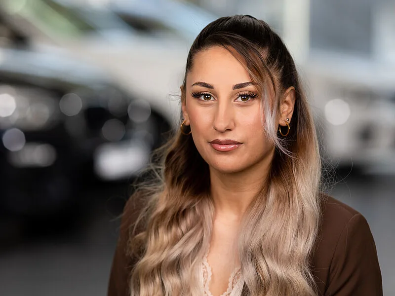 Kaltenbach Gruppe – Eine junge Frau mit langen, gewellten blonden Haaren und Ohrringen schaut mit neutralem Blick in die Kamera. Sie trägt eine braune Jacke und ein cremefarbenes Oberteil. Im Hintergrund sind unscharfe Autos in der Nähe von Kaltenbach-Remscheid zu sehen. – BMW, MINI, KIA, BMW Motorrad & Honda Motorrad Partner in NRW