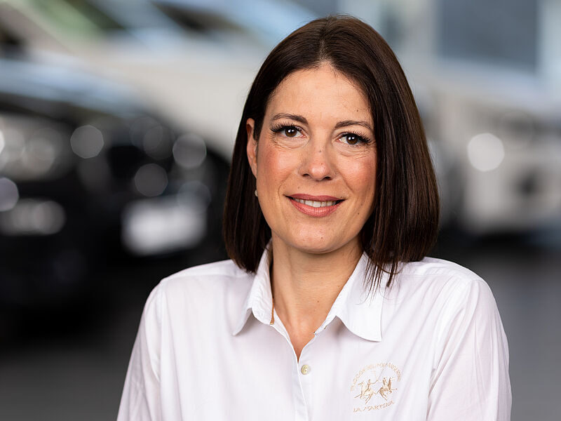 Kaltenbach Gruppe – Eine Frau mit glattem braunem Haar und weißem Kragenhemd lächelt in die Kamera. Im unscharfen Hintergrund, der sich wahrscheinlich in Kaltenbach-Remscheid befindet, sind scheinbar Autos in Innenräumen zu sehen. – BMW, MINI, KIA, BMW Motorrad & Honda Motorrad Partner in NRW