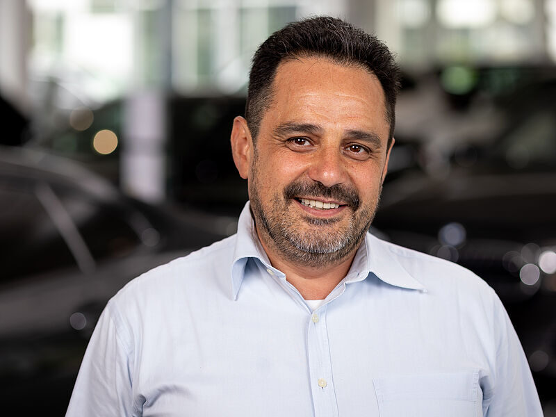 Kaltenbach Gruppe – Ein Mann mittleren Alters mit kurzen dunklen Haaren und einem Bart, der ein hellblaues Hemd mit Kragen trägt, lächelt in einer hellen Halle in Kaltenbach-Wiehl in die Kamera, während im Hintergrund Autos verschwimmen. – BMW, MINI, KIA, BMW Motorrad & Honda Motorrad Partner in NRW