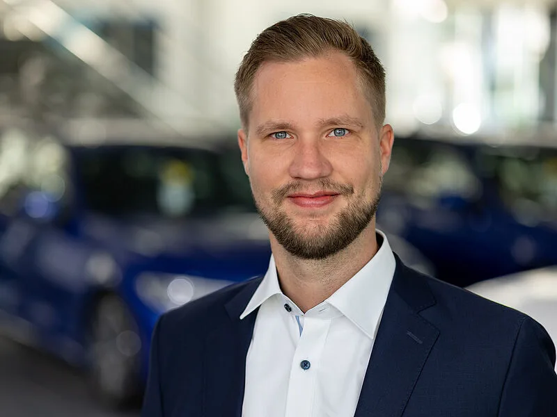 Kaltenbach Gruppe – Ein Mann mit kurzem blondem Haar und gestutztem Bart, der eine dunkelblaue Anzugsjacke und ein weißes Hemd trägt, lächelt in die Kamera. Im Hintergrund sind unscharfe blaue Autos zu sehen, die auf einen überdachten Ausstellungsraum in Kaltenbach Remscheid hindeuten. – BMW, MINI, KIA, BMW Motorrad & Honda Motorrad Partner in NRW